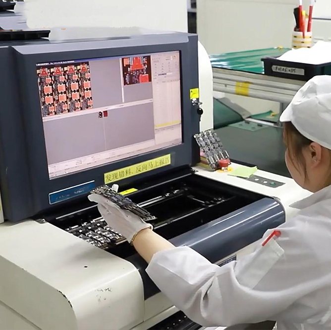 Operator loading a board into an automated optical inspection machine, with the inspection images shown on the screen